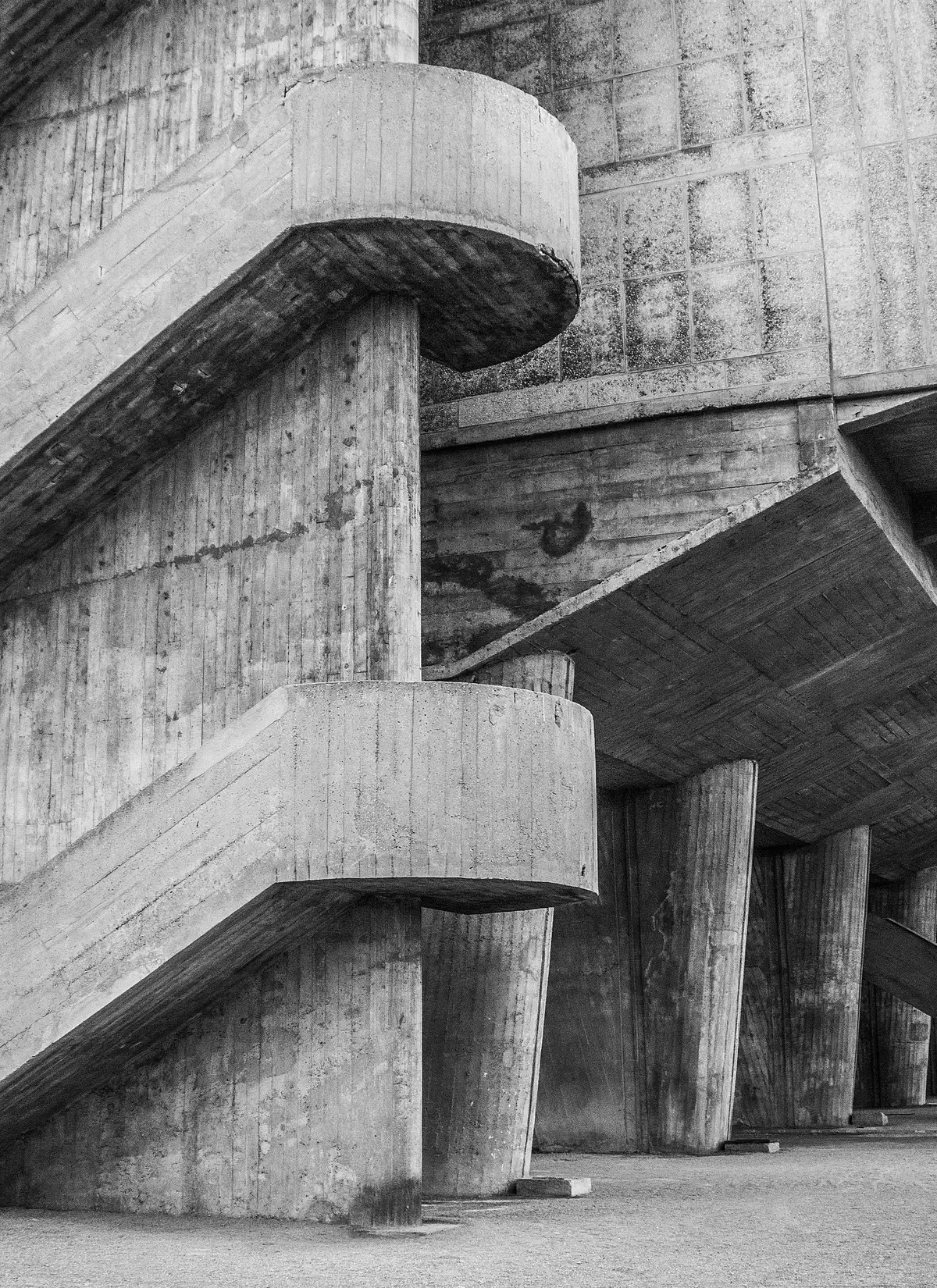 A brutalist concrete stair: cantilevered cylindrical landings above board-marked formwork, pilotis receding into shadow.